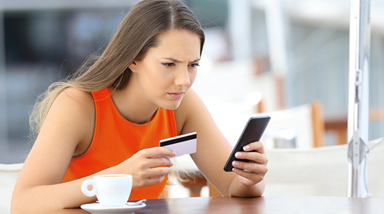 Woman at a coffee table staring intently at her smartphone in one hand while holding a bank card in the other.