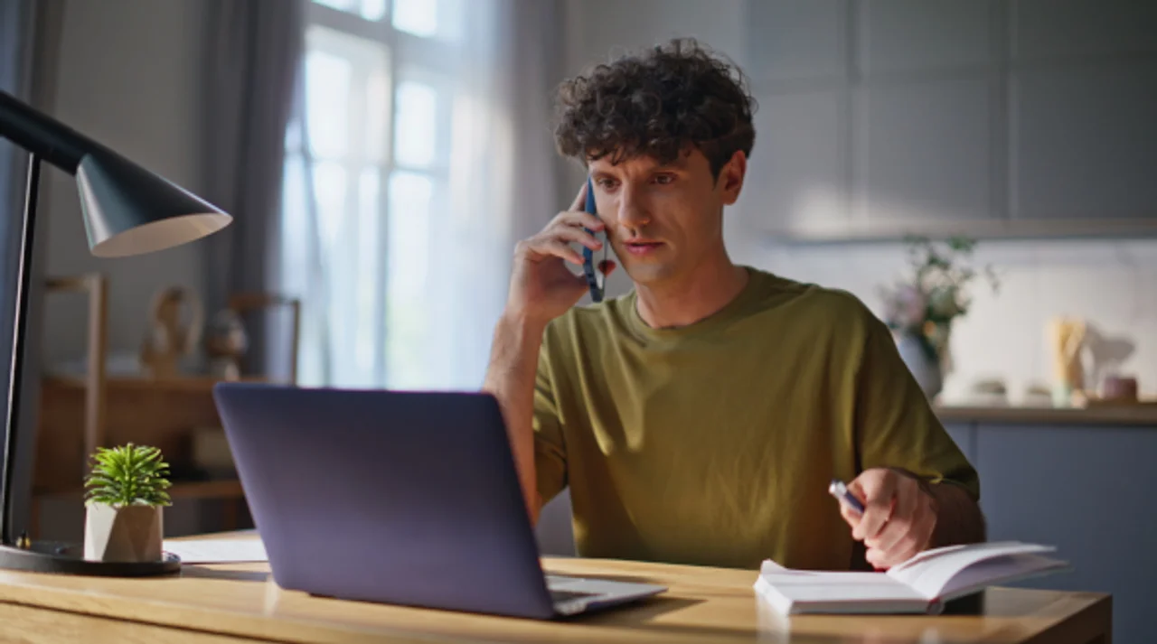 Ein Mann sitzt am Küchentisch und telefoniert, während er auf seinen aufgeklappten Laptop blickt.
