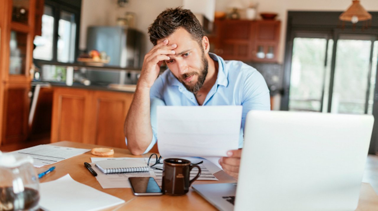 Stressed man sits at a kitchen table in front of a laptop, holding a document and resting his forehead on his other hand.