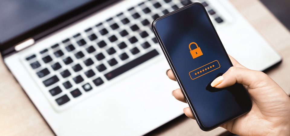 Eine Hand, die ein Smartphone mit einem Schlosssymbol und einem anonymisierten PIN zeigt, dahinter ein aufgeklappter Laptop