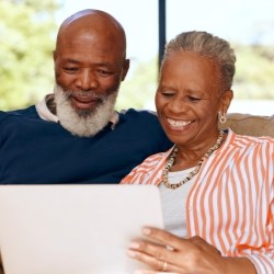 Two elderly people sitting on a sofa, smiling as they look at a tablet